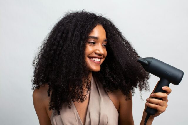 Een lachende vrouw met krullen houdt een föhn bij haar haar