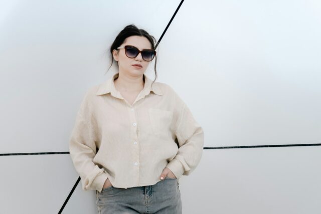 Een vrouw in een beige overshirt met zonnebril staat voor een lichte muur